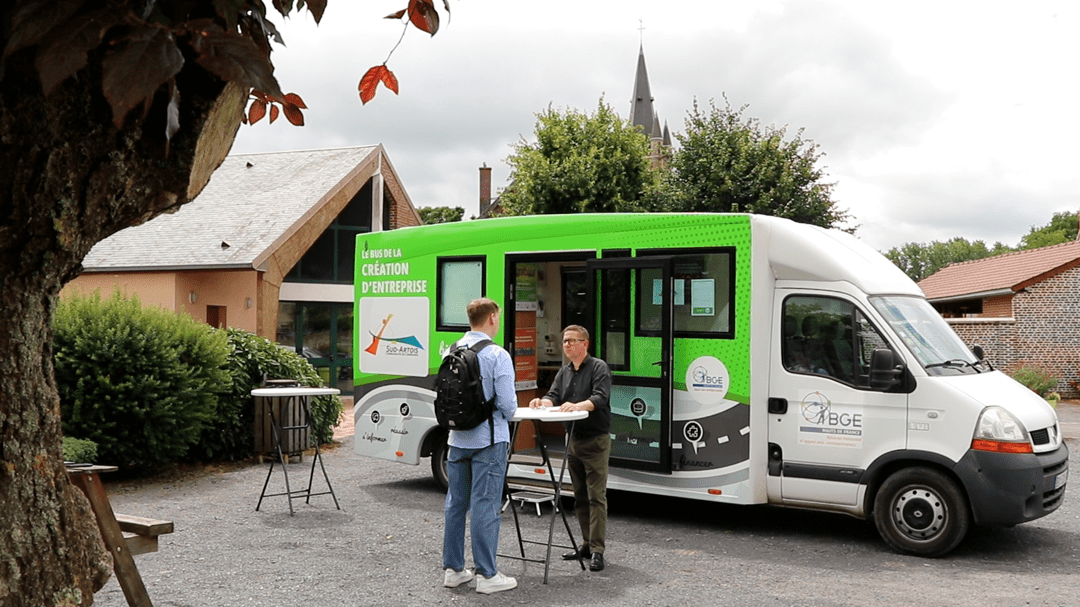 Un conseiller discute avec un habitant devant le bus de la création d'entreprise sur la place d'un village des Hauts-de-France.