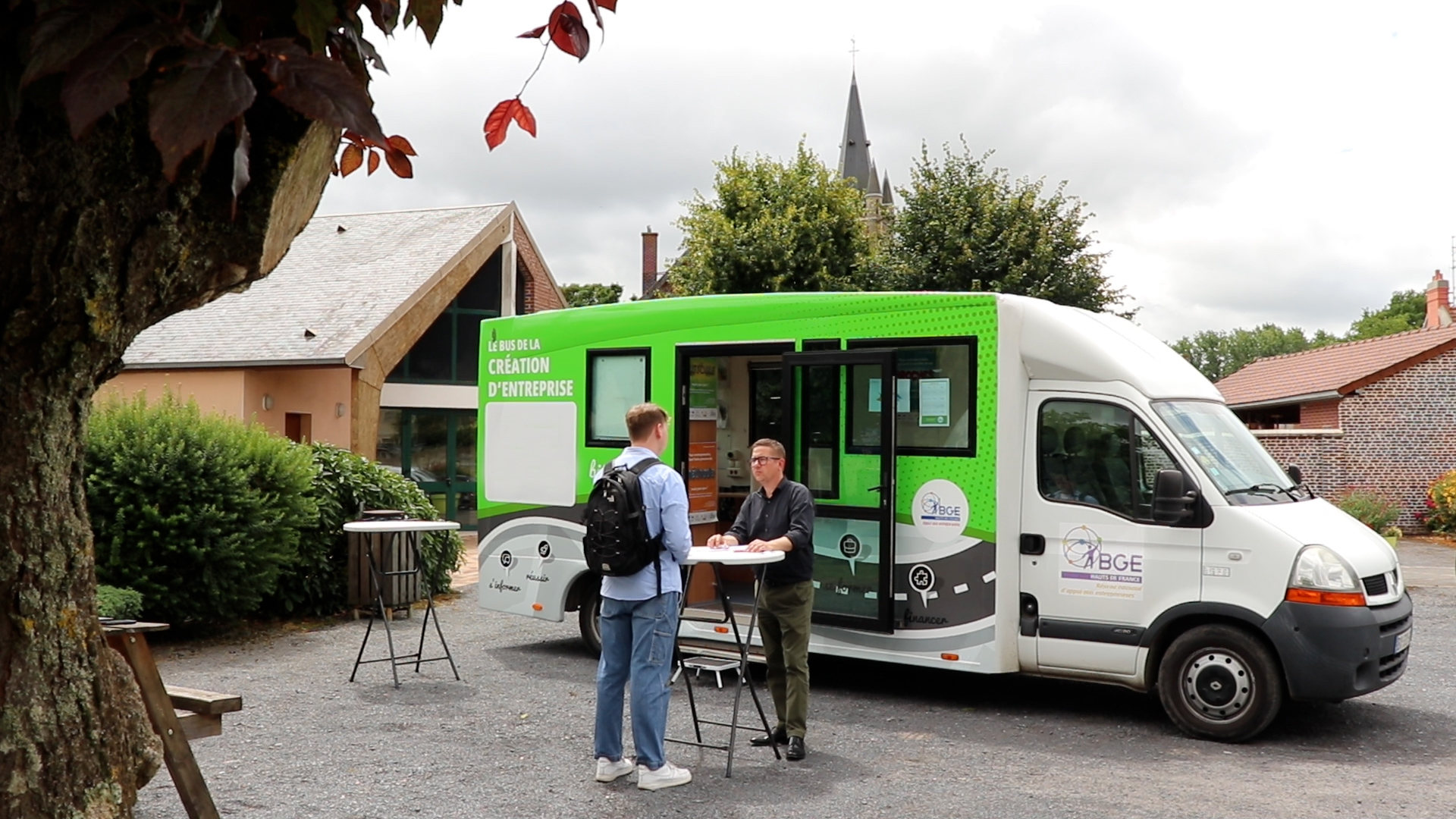 Un conseiller discute avec un habitant devant le bus de la création d'entreprise sur la place d'un village des Hauts-de-France.